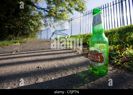 Leere Bierflasche der Marke Stella Artois auf dem Bürgersteig mit einem verlassenen Einkaufswagen im Hintergrund Stockfoto