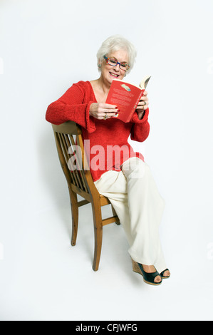Attraktive ältere Dame in helle Kleidung saß auf Stuhl Buch genommen vor einem weißen Hintergrund Stockfoto