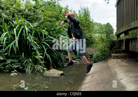 8 Jahre alten kaukasischen junge springt auf Felsen über Stream auf Barrs Gerichts graben Teich in der Nähe von Longwell Green in Bristol, Großbritannien Stockfoto