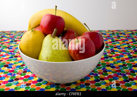 Obstschale sitzen in einer weißen Schüssel auf einem bunten Hintergrund. Banane, Birne, Apfel und Zitronen Stockfoto