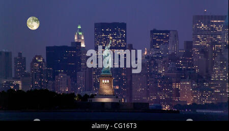 Ein Vollmond steigt in Lower Manhattan, New York City, in dieser Ansicht von Bayonne, New Jersey. Im Mittelpunkt steht die Statue of Liberty. Links von der Mitte ist das Woolworth Building; rechts von der Mitte ist das goldene Wappen der NY City Municipal Building. Stockfoto