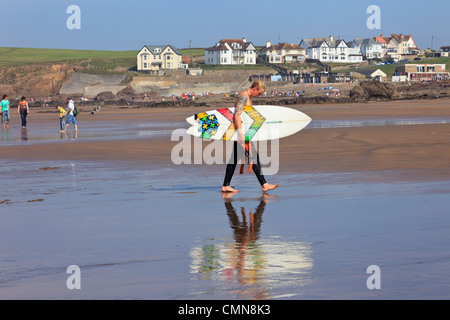 Surfer tragen ein Surfbrett über Crooklets Strand mit Reflexionen im nassen Sand in Bude, North Cornwall, England, UK, Großbritannien Stockfoto