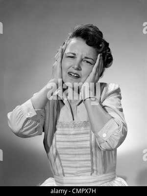 1950ER JAHREN FRAU IN BAUMWOLL-KLEID MIT BEIDEN HÄNDEN ÜBER IHRE OHREN HIELT IHR STIRNRUNZELND KOPF Stockfoto