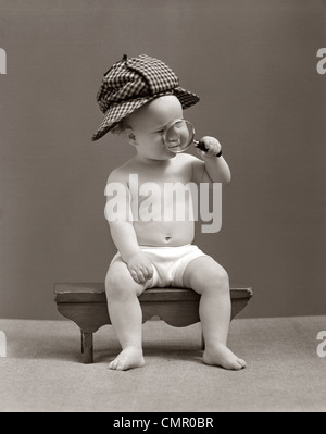 1940ER JAHRE BABY SHERLOCK HOLMES IN WINDEL SITZEN AUF BANK TRAGEN DEER STALKER HUT BLICK DURCH DIE LUPE Stockfoto