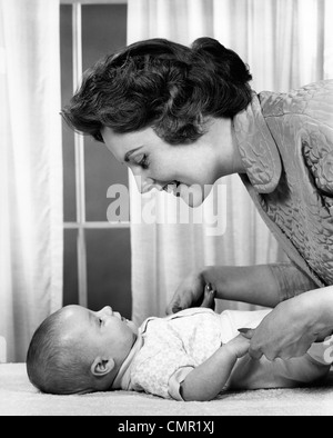 1950S 1960S MUTTER LÄCHELTE BABY LIEGEND AUF TISCH INDOOR Stockfoto