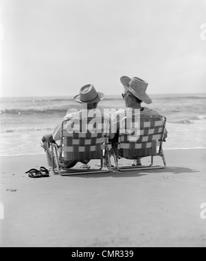 RÜCKANSICHT DER 1970ER JAHRE ÄLTERES EHEPAAR IN STROHHÜTEN SITZEN AM STRAND STÜHLE AM UFER Stockfoto