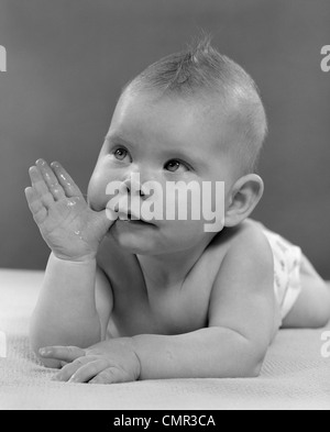 1950ER JAHRE PORTRAIT BABY LIEGEND AUF BAUCH MIT DAUMEN IM MUND Stockfoto