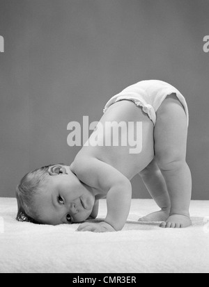 1950ER JAHREN SEITENANSICHT PORTRÄT BABY WINDEL MIT WANGE AN BODEN UND UNTEN IN LUFT BLICK IN DIE KAMERA Stockfoto