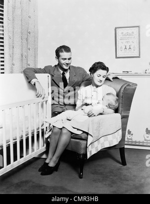 1930S 1940S MUTTER SAß IM STUHL IM KINDERGARTEN BABY 11 MONATE AUF RUNDE TRINKT AUS FLASCHE VATER STÜTZTE SICH AUF KRIPPE AUF Stockfoto