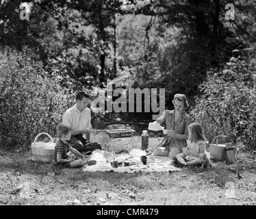 1940ER JAHRE FAMILIE MUTTER VATER SOHN TOCHTER PICKNICKEN MIT BARBECUE GRILL IM PARK Stockfoto