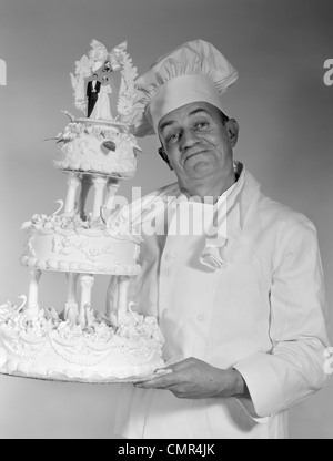 1950S 1960S ÄLTEREN MANN CHEF BLICK IN DIE KAMERA LÄCHELN HOLDING 3-TIER-HOCHZEITSTORTE Stockfoto