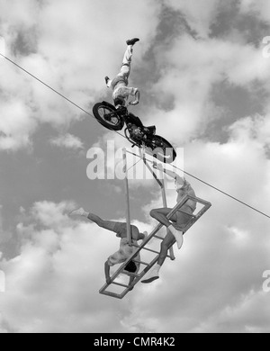 1950S 1960S HOCHSEILAKT MIT AUF DEM KOPF STEHENDEN MANN AUF DEM MOTORRAD MIT MANN UND FRAU AUF AUSGEWOGENE LEITER UNTEN AUSGESETZT Stockfoto