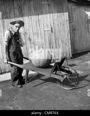 1970S 1980S ÄLTERE LANDWIRT IN OVERALLS & STROHHUT BLICK IN DIE KAMERA, DIE ALTE SCHUBKARRE MIT RIESIGEN KÜRBIS DRAUF DRÜCKEN Stockfoto