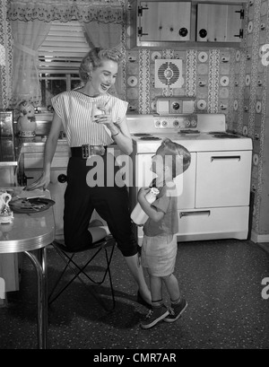 1950ER JAHRE JUNGE IN KÜCHE HOLDING MILCHFLASCHE BLICKTE ZU MUTTER TRINKGLAS MILCH Stockfoto