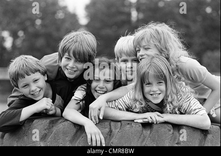 1970S 1980S GRUPPE VON SECHS JUNGEN & MÄDCHEN VERSAMMELTEN SICH AM GROßEN SPIELPLATZ REIFEN Stockfoto