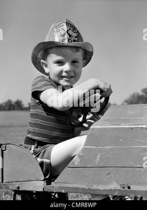 1940ER JAHRE JUNGEN SPIELEN FEUERWEHRMANN IN SPIELZEUG FEUER LKW TRAGEN FIREMAN ES SICHERHEIT HUT HÄLT LENKRAD LÄCHELND Stockfoto