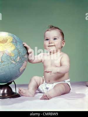 1960ER JAHREN LÄCHELN BABYMÄDCHEN SITZEN IN WINDELN HAND BERÜHREN WELTKUGEL Stockfoto