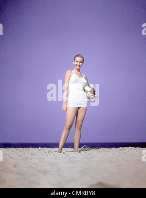 1950ER JAHRE VOLLER LÄNGE PORTRAIT FRAU LEICHTEN BLAUEN EINTEILER BADEANZUG SCHWIMMEN TRAGEN AUF SAND HOLDING STRANDMODE BALL STEHEND Stockfoto