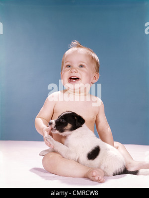 1960ER JAHRE LÄCHELN BABYMÄDCHEN IN WINDELN MIT JUNGEN SCHWARZ / WEIß-HÜNDCHEN SPIELEN SITZEN Stockfoto