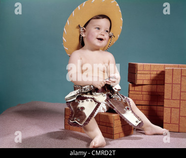 1960ER JAHREN LÄCHELN BABY TRÄGT COWBOYHUT UND SPIELZEUGPISTOLEN UND WESTLICHEN HOLSTER Stockfoto