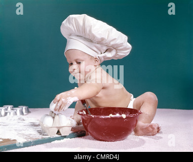 1960ER JAHRE BABY TRAGEN KÖCHE HAT HOLDING EI UMGEBEN VON KOCHAUSRÜSTUNG Stockfoto