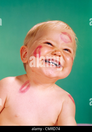 1960ER JAHRE PORTRÄT DES LÄCHELNDEN GLÜCKLICHES BABY MIT ROTEM LIPPENSTIFT KÜSSEN SPUREN IM GESICHT Stockfoto