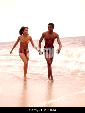 1970ER JAHREN PAAR AFROAMERIKANISCHE JUNGEN TRAGEN BADEANZÜGE IN MEERESBRANDUNG AUSGEFÜHRT Stockfoto