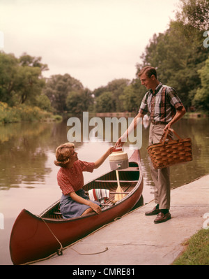 1960ER JAHRE JUNGES PAAR LADEN PICKNICK KORB THERMOSKANNE IN KANU Stockfoto