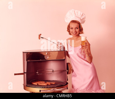 1960ER JAHREN LÄCHELND BLONDE FRAU TRÄGT ROSA KLEID UND KOCH HUT GRILLEN HEIß HUNDE BIER TRINKEN Stockfoto