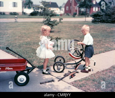 KLEINES MÄDCHEN HÄLT PUPPE LITTLE BOY REPARATUR FAHRRAD MIT WERKZEUG IM FREIEN BEOBACHTEN Stockfoto