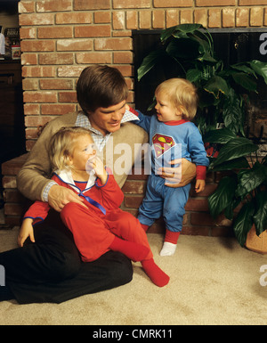 1980ER JAHREN VATER SITZEN KAMIN MIT ZWEI KLEINEN KINDERN IM PYJAMA Stockfoto