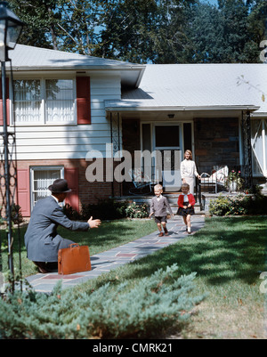 1950ER JAHREN VATER HEIMKEHREN KNIEND ARME AUSGESTRECKT JUNGE MÄDCHEN LÄUFT UM IHN FRAU STAND VORSTADTHAUS FAMILIE RETRO-WILLKOMMEN Stockfoto