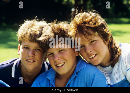 1980ER JAHREN ZWEI BRÜDER UND IHRE SCHWESTER STELLTE KÖPFE ZUSAMMEN LÄCHELNDEN BLICK IN DIE KAMERA Stockfoto