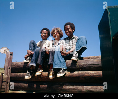 1970ER JAHRE GLOCKE 3 JUNGEN SIT LOG ZAUN LÄCHELN AUF DEN LIPPEN TRAGEN BLAUE AUSGESTELLTEM BEIN JEANS UNTEN 2 JUNGEN AFRO-AMERIKANISCHEN 1 CAUCASION RASSISCHE MISCHUNG Stockfoto