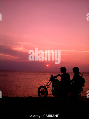 1970 1970S SILHOUETTE PÄRCHEN SONNENUNTERGANG STRAND REITEN CHOPPER MOTORRAD MOTORRAD MOTORRAD MOTORRAD BIKER BIKES Stockfoto