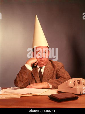 1960ER JAHREN TRAURIG DEPRIMIERT GESCHÄFTSMANN SCHREIBTISCH TRAGEN DUNCE CAP MANN MÄNNER HANDELSLEHRER FEHLER FEHLER FALSCHE LUSTIGE SILLY Stockfoto