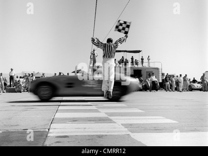 1960ER JAHRE MANN SPRINGEN WINKEN CHECKERED FLAG AS SPORTWAGEN ÜBER DIE ZIELLINIE ZU GEWINNEN Stockfoto