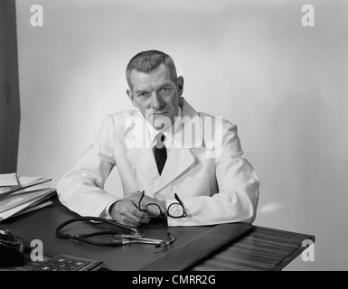 1950S 1960S PORTRAIT MÄNNLICHEN ARZT SITZEN AM SCHREIBTISCH ERNSTEN AUSDRUCK Stockfoto