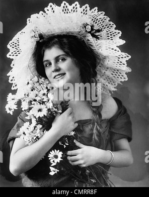 1900S 1910S UMDREHUNG DER JAHRHUNDERT-FRAU MIT LANGEN ZÖPFEN, AUSGEFALLENE SPITZEN HUT & HÄLT BLUMENSTRAUSS SONNENHUT Stockfoto