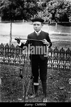 1800 S-1890 S-1900 S WENDE DES JAHRHUNDERTS JUNGEN STEHEN IM HOF NEBEN ALTEN ALTMODISCHEN FAHRRAD Stockfoto