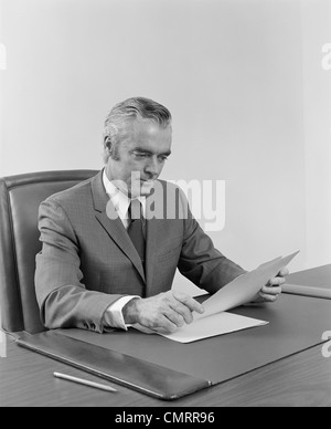 1970S 1960S IM ALTER GESCHÄFTSMANN EXECUTIVE MITTE SITZEN AM SCHREIBTISCH LESEN PAPIERE Stockfoto