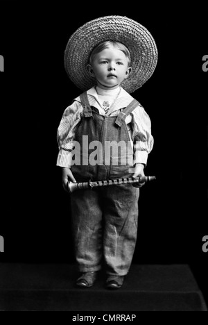 1800S 1890S UMDREHUNG DES JAHRHUNDERTS JUNGEN IN OVERALLS & STROHHUT HOLDING KLEINE KLARINETTE-ÄHNLICHES INSTRUMENT Stockfoto