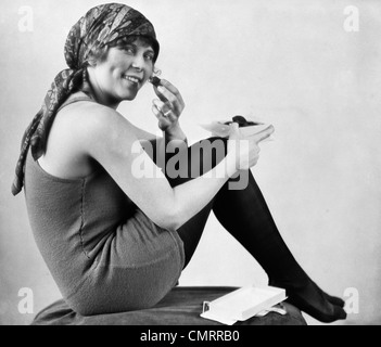 1920ER JAHREN FRAU IN ALTEN ALTMODISCHEN BADEANZUG MIT SCHAL AUF KOPF SITZEND MIT KNIE ZUR BRUST ESSEN SCHOKOLADE BLICK IN DIE KAMERA Stockfoto