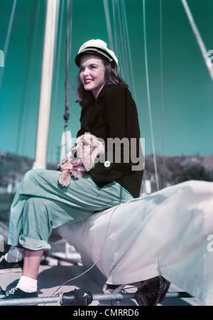 1940S 1950S LÄCHELNDE FRAU MIT SEGELN YACHTING OUTFIT SITZEN AM RAND DES SEGELBOOT HOLDING WELPEN IN RUNDE Stockfoto