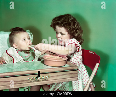 1940S 1950S MÄDCHEN SITZEN FÜTTERUNG BABYSCHWESTER IN HOHER STUHL Stockfoto