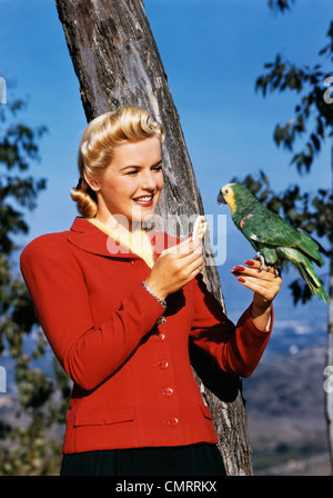 1940ER JAHRE LÄCHELNDEN BLONDEN FRAU FÜTTERUNG CRACKER PAPAGEI IM FREIEN Stockfoto