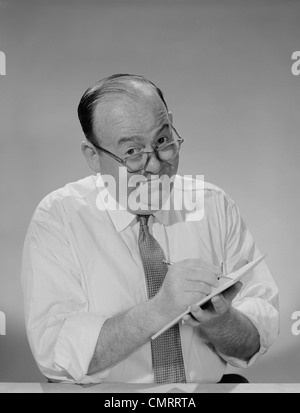 1950S 1960S ÄRMEL GLATZENBILDUNG GESCHÄFTSMANN TRAGEN BRILLEN HEMD LÄCHELND SCHREIBEN NOTEPAD BLICK IN DIE KAMERA Stockfoto