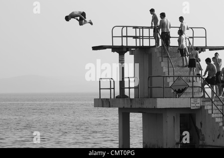 Ein Schuss von einem Mann somersaulting aus blackrock Sprungbrett, während eine Gruppe von Schaulustigen in Badekleidung aus den Schritten beobachten. Stockfoto