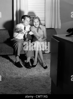 1950ER JAHREN PAAR IM ABGEDUNKELTEN WOHNZIMMER FERNSEHEN MIT MANN ZEIGTE AUF BILDSCHIRM Stockfoto