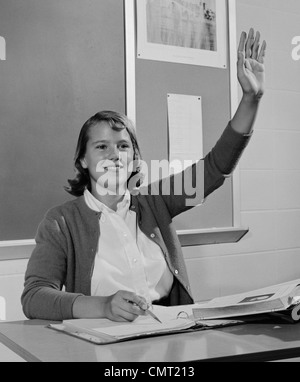 1960ER JAHREN LÄCHELND TEEN MÄDCHEN SITZEN KLASSENZIMMER SCHREIBTISCH HEBEN IHRE HAND Stockfoto